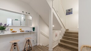 Una cocina moderna con encimera blanca, dos taburetes, una planta colgante y una escalera con escalones de madera.