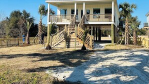 Una casa frente al mar de dos pisos, con balcón, escaleras de madera y camino de entrada de arena.