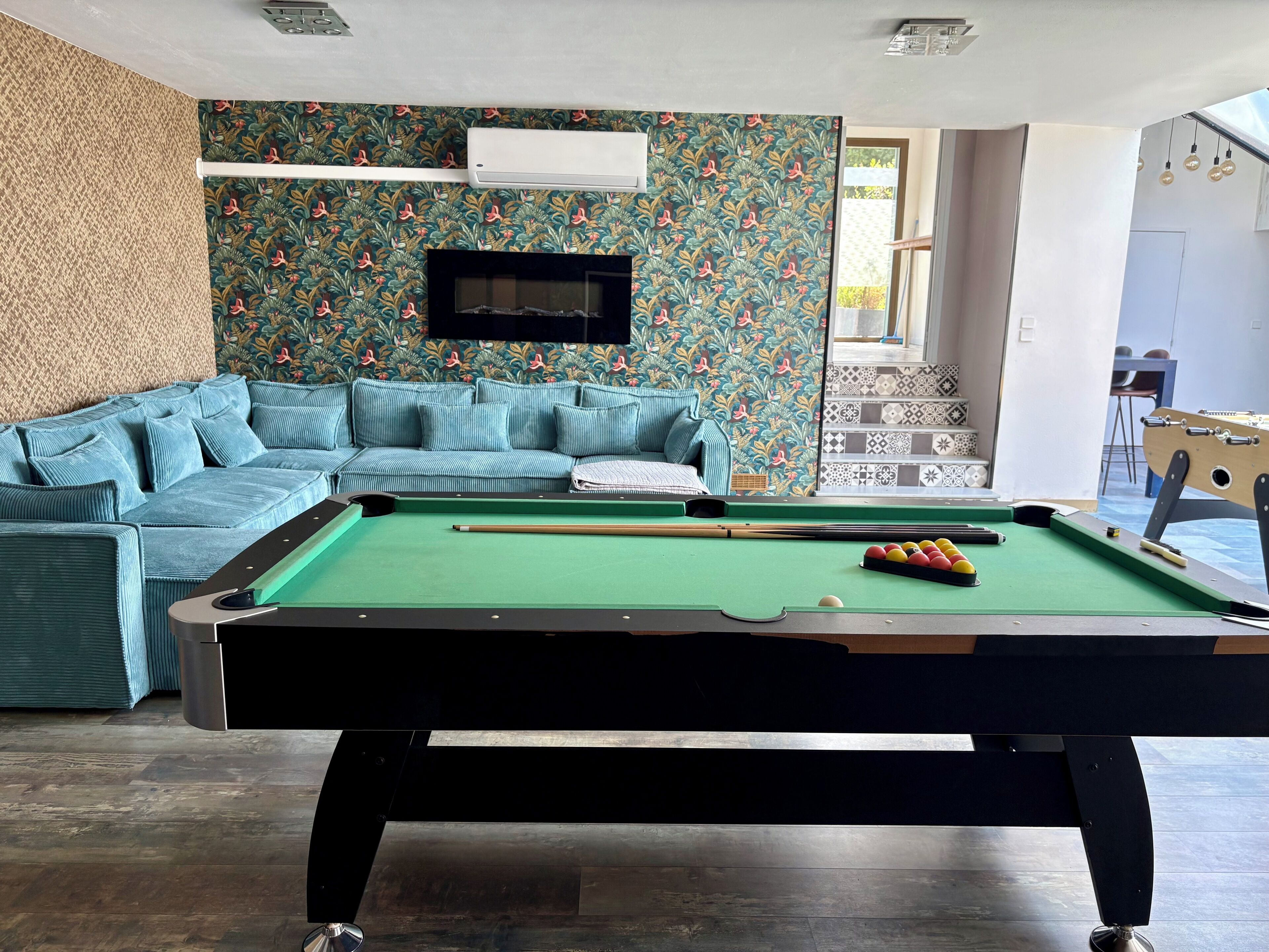 A pool table with balls, a sofa, and a television in a room with patterned wallpaper.