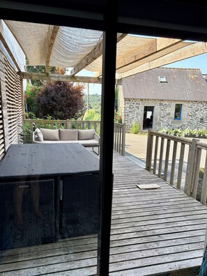 Eine Holzterrasse mit einer Rattan-Couch und einem Tisch, im Hintergrund ein Steinhaus und Blick ins Grüne.