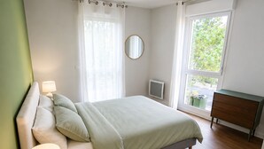 A bedroom with a bed, bedside lamps, a dresser, a mirror, and a window with a view of greenery.