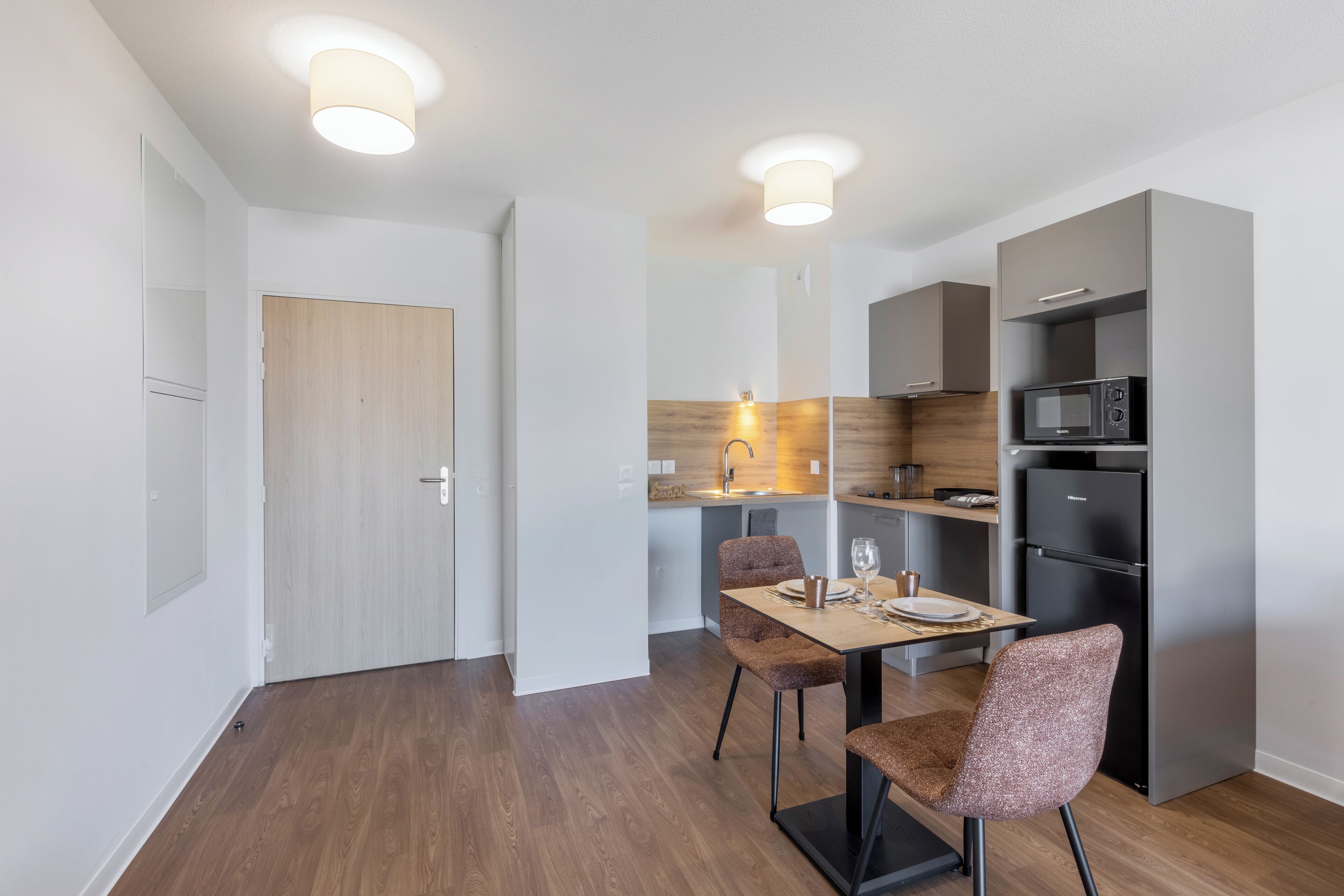 A modern kitchen with a microwave, refrigerator, and wooden cabinets. There is a dining area with two chairs and a table.