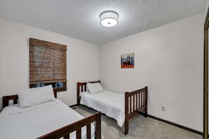 A room with two wooden beds, a window with blinds, a ceiling light, and a small wall-mounted picture.
