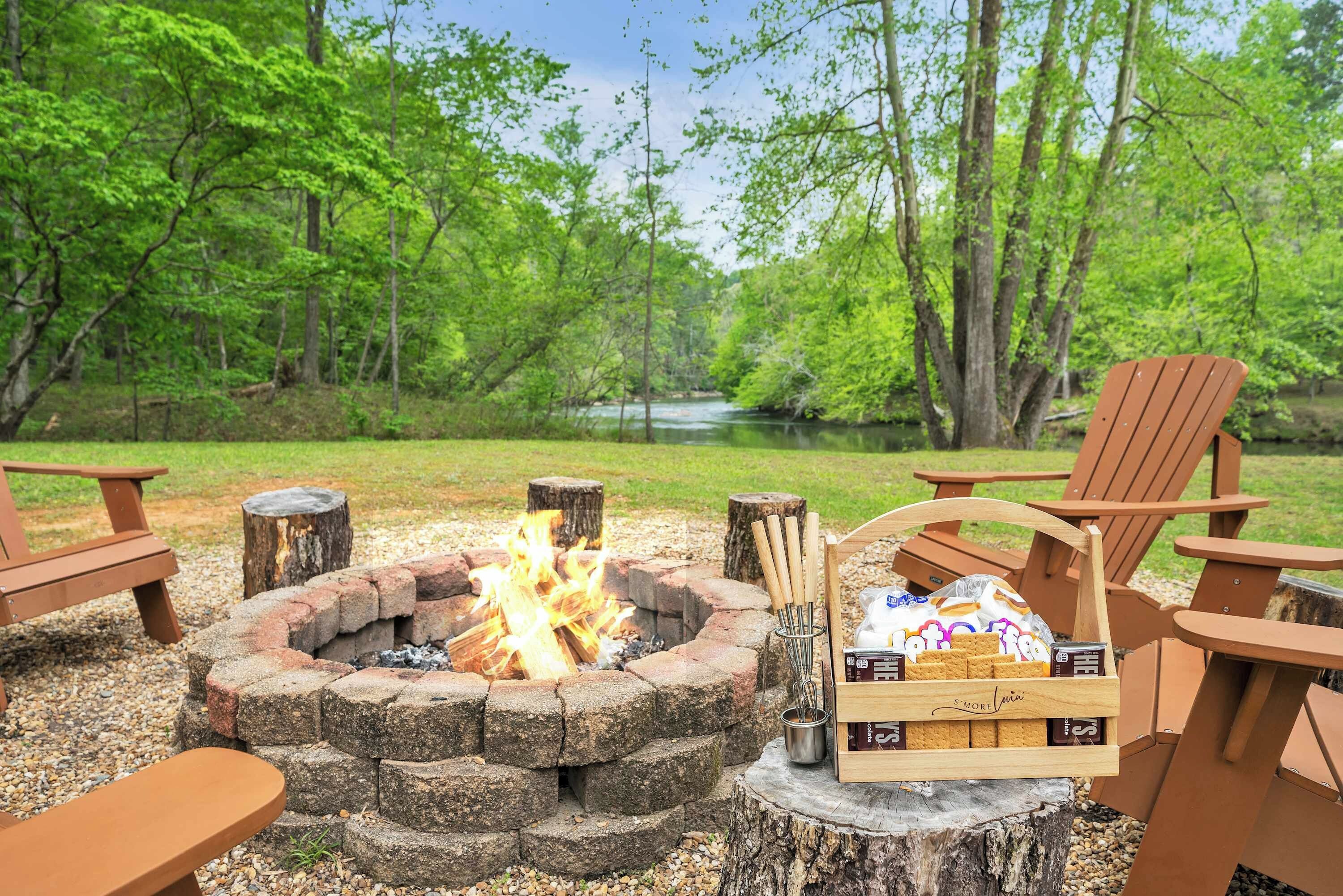 Un feu de camp entouré de chaises en bois et d’une table de pique-nique, installé dans une zone boisée avec un lac en arrière-plan.
