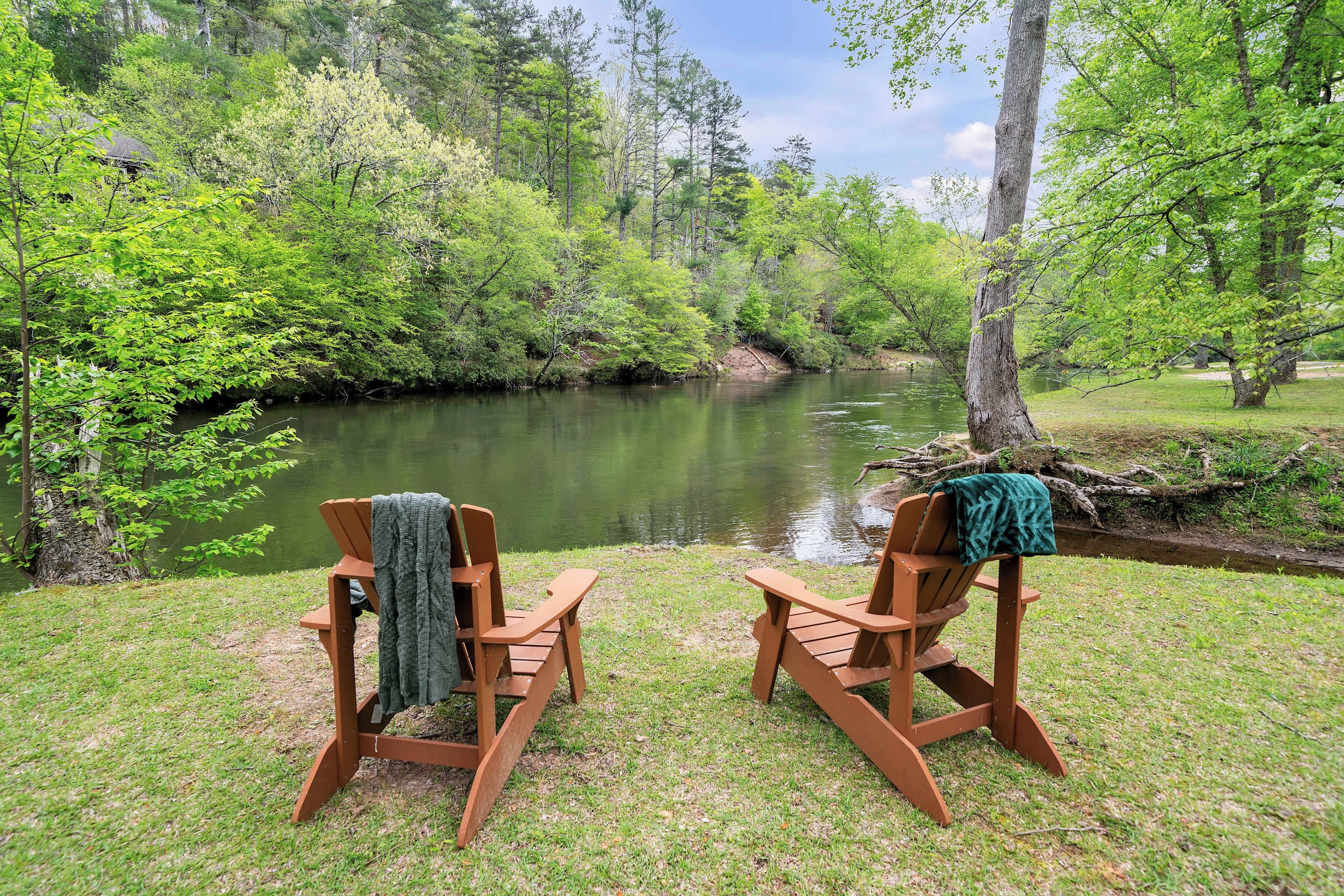 Two wooden Adirondack chairs facing a river with lush greenery and trees in the background.