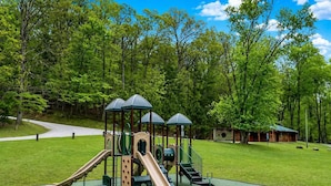 A playground with slides and climbing structures, surrounded by a grassy area and trees.