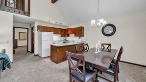 A dining area with a wooden table and chairs, a kitchen with wooden cabinets, a clock on the wall, and a staircase leading to an upper level.