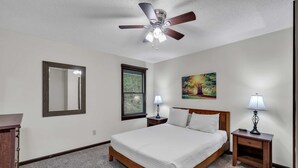 A bedroom with a bed, a ceiling fan, a nightstand with a lamp, a bedside table, a window with blinds, and a painting on the wall.