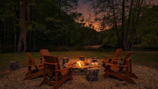 A campfire with wooden chairs arranged around it in a forest setting.