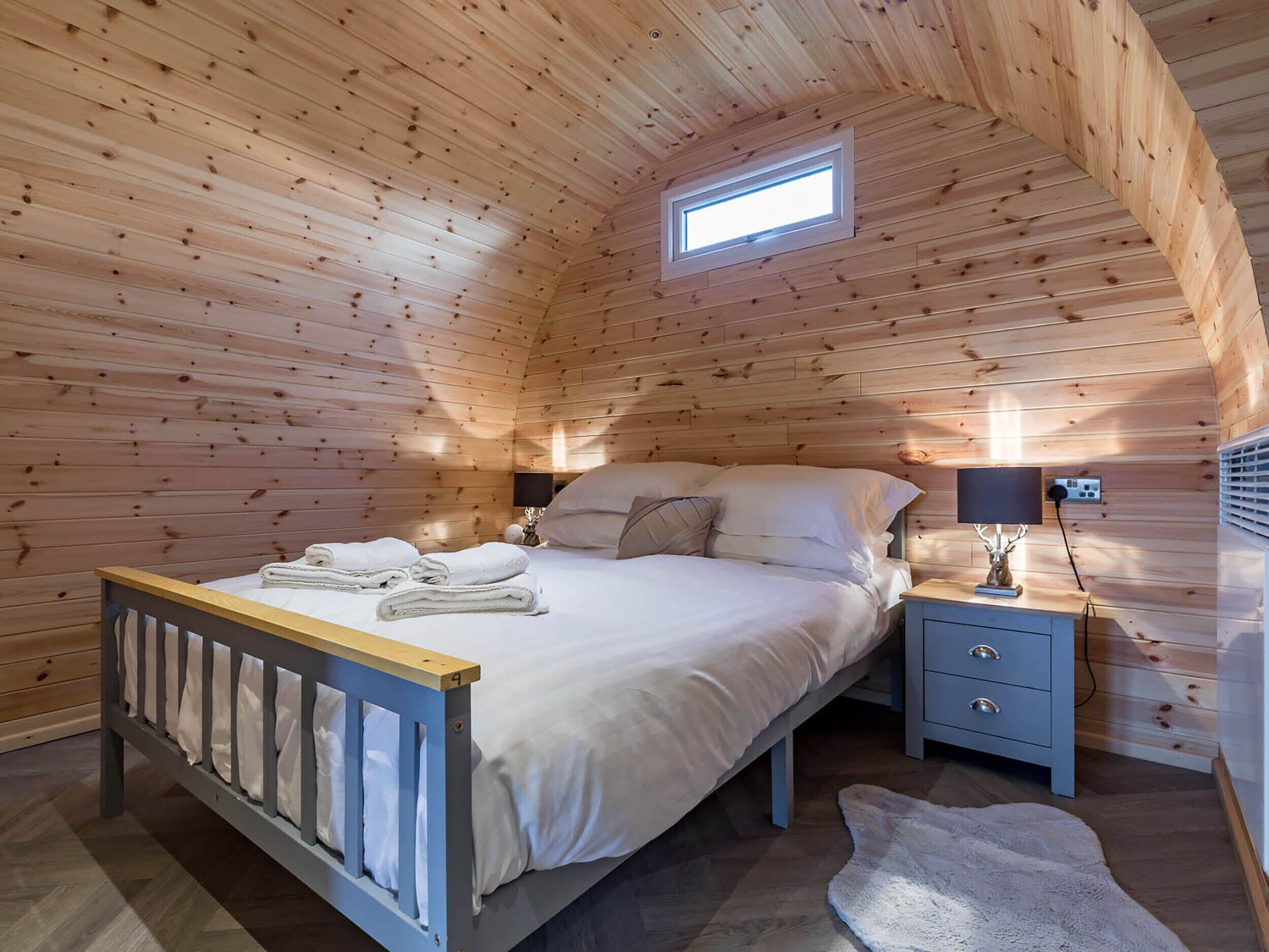 A wooden cabin bedroom with a bed, nightstands, a lamp, and a small window.