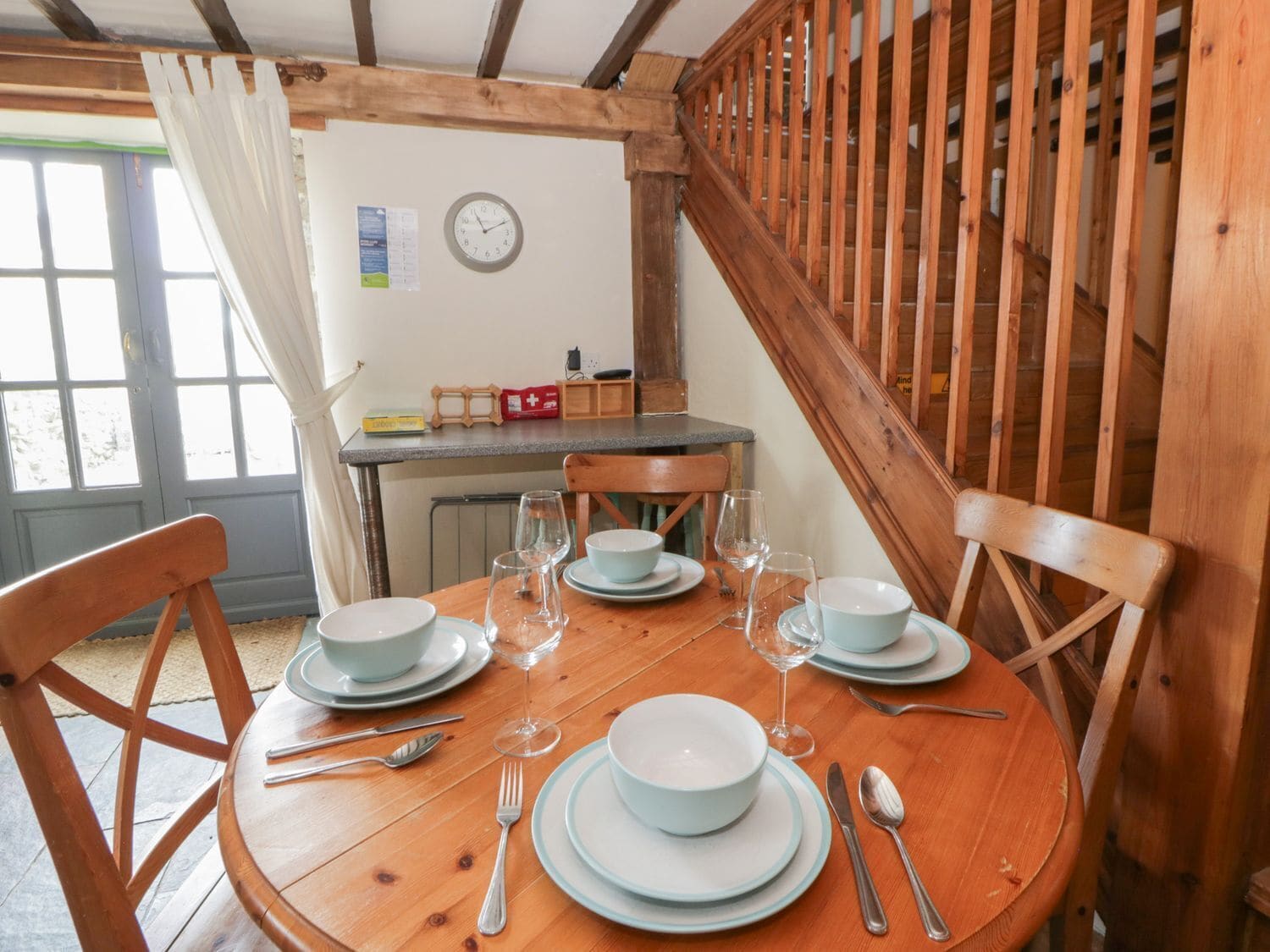 Una mesa de comedor de madera dispuesta con platos, cuencos, cubiertos y copas de vino en una habitación con vigas de madera y un reloj en la pared.