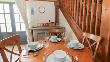 Una mesa de comedor de madera dispuesta con platos, cuencos, cubiertos y copas de vino en una habitación con vigas de madera y un reloj en la pared.