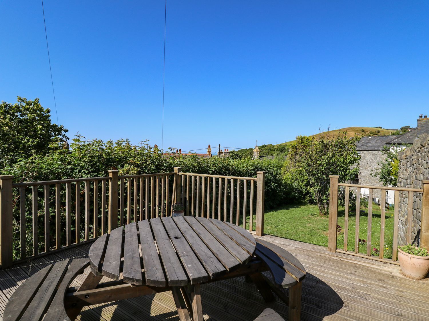 A wooden deck with a round table and benches, surrounded by a wooden fence, overlooking a green garden and a stone building.