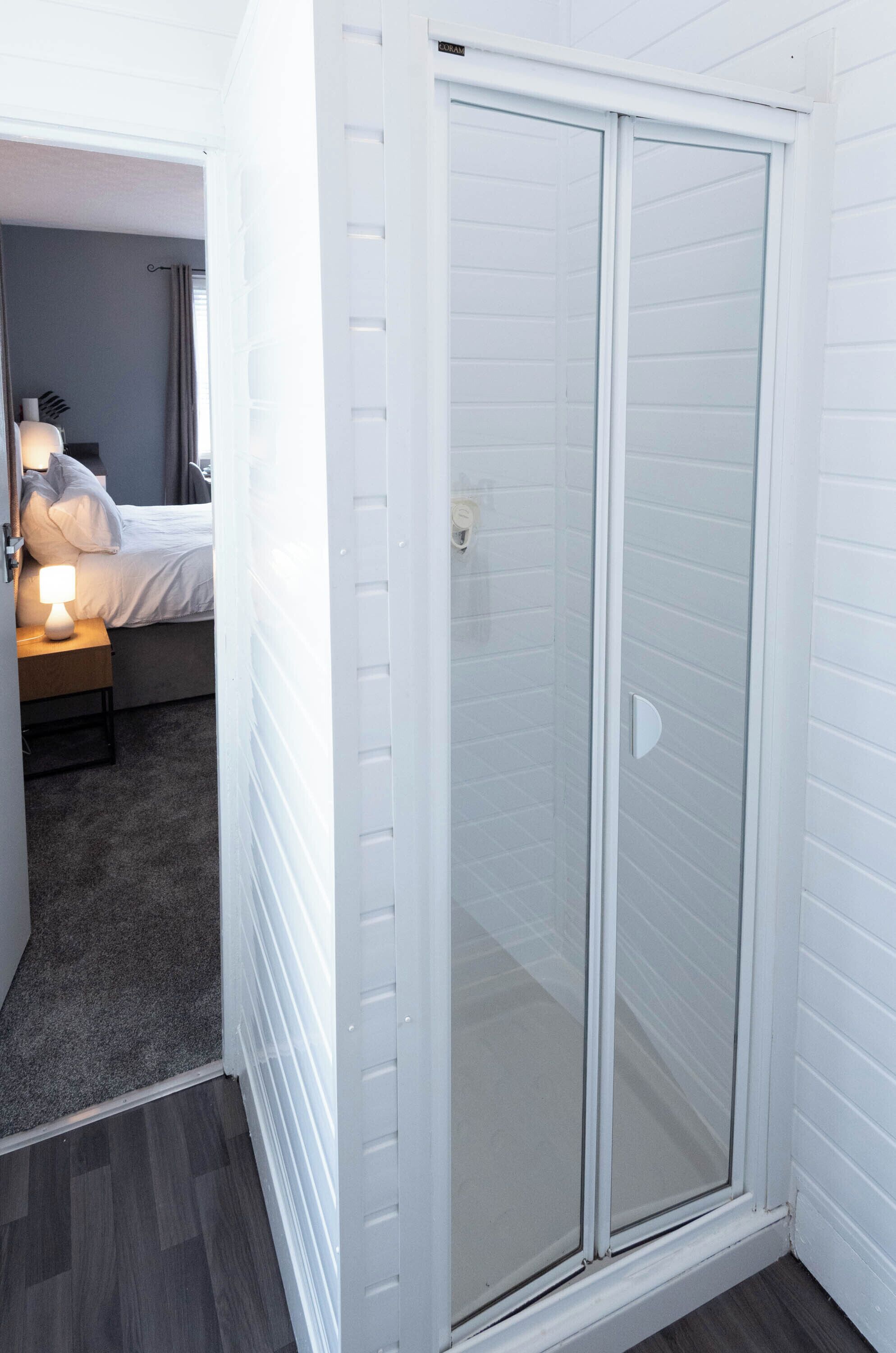 A room with a glass-enclosed shower, a bed with white bedding, and a nightstand with a lamp.
