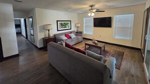 Un salon avec un canapé gris, une table basse et un ventilateur de plafond. Un tableau encadré est accroché au mur, et il y a une fenêtre avec des stores.