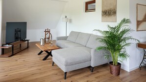 Ein Wohnzimmer mit einem grauen Ecksofa, einem Holztisch, einer Stehlampe und einem Blumentopf.