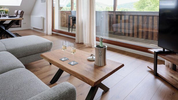 Ein modernes Wohnzimmer mit Holzboden, grauem Sofa, einem Couchtisch mit Gläsern und einer Flasche sowie einem Fernseher auf einem Holzbodenständer.
