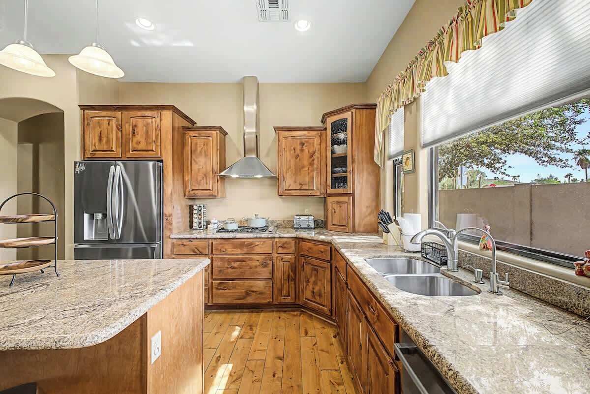 A kitchen with wooden cabinets, a stainless steel refrigerator, granite countertops, a built-in oven, and a window with a view.