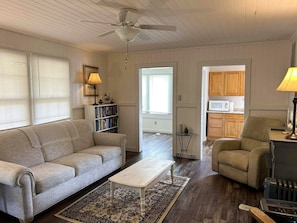 Ein Wohnzimmer mit Sofa, Couchtisch und Bücherregal. Es gibt einen Deckenventilator, eine Tür zu einer Küche und ein Fenster mit Jalousien.
