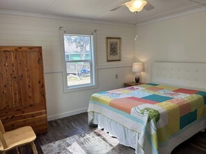 A bedroom with a wooden wardrobe, a window with a view, a bed with a colorful quilt, a bedside lamp, and a ceiling fan. - IR54: Cedarberry | Conveniently Located in the Heart of the Village (Ocracoke)