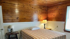 Una habitación con paneles de madera, una cama, una mesita de noche con una lámpara y una ventana.