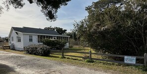A white single-story house with a dark roof, surrounded by trees and a wooden fence. - IR54: Cedarberry | Conveniently Located in the Heart of the Village (Ocracoke)