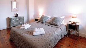 Un lit bien fait, recouvert d’une couverture grise, avec deux lampes de chevet et une commode à tiroirs.