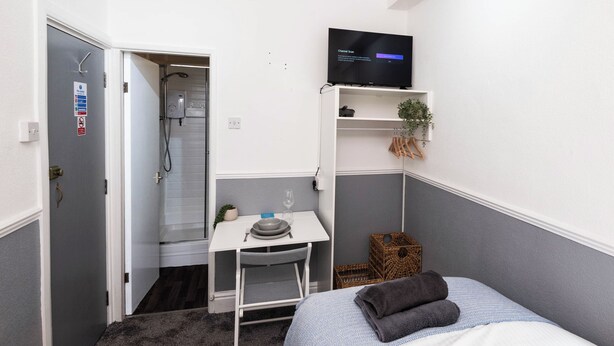 A compact hotel room with a shower, a small desk with a chair, a bed, a TV, and a plant. - The Bay Apart-Hotel by Guestz (Morecambe)