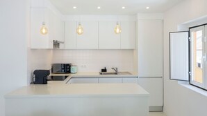 Une cuisine moderne avec des meubles blancs, un four encastré, une machine à café et des luminaires suspendus.