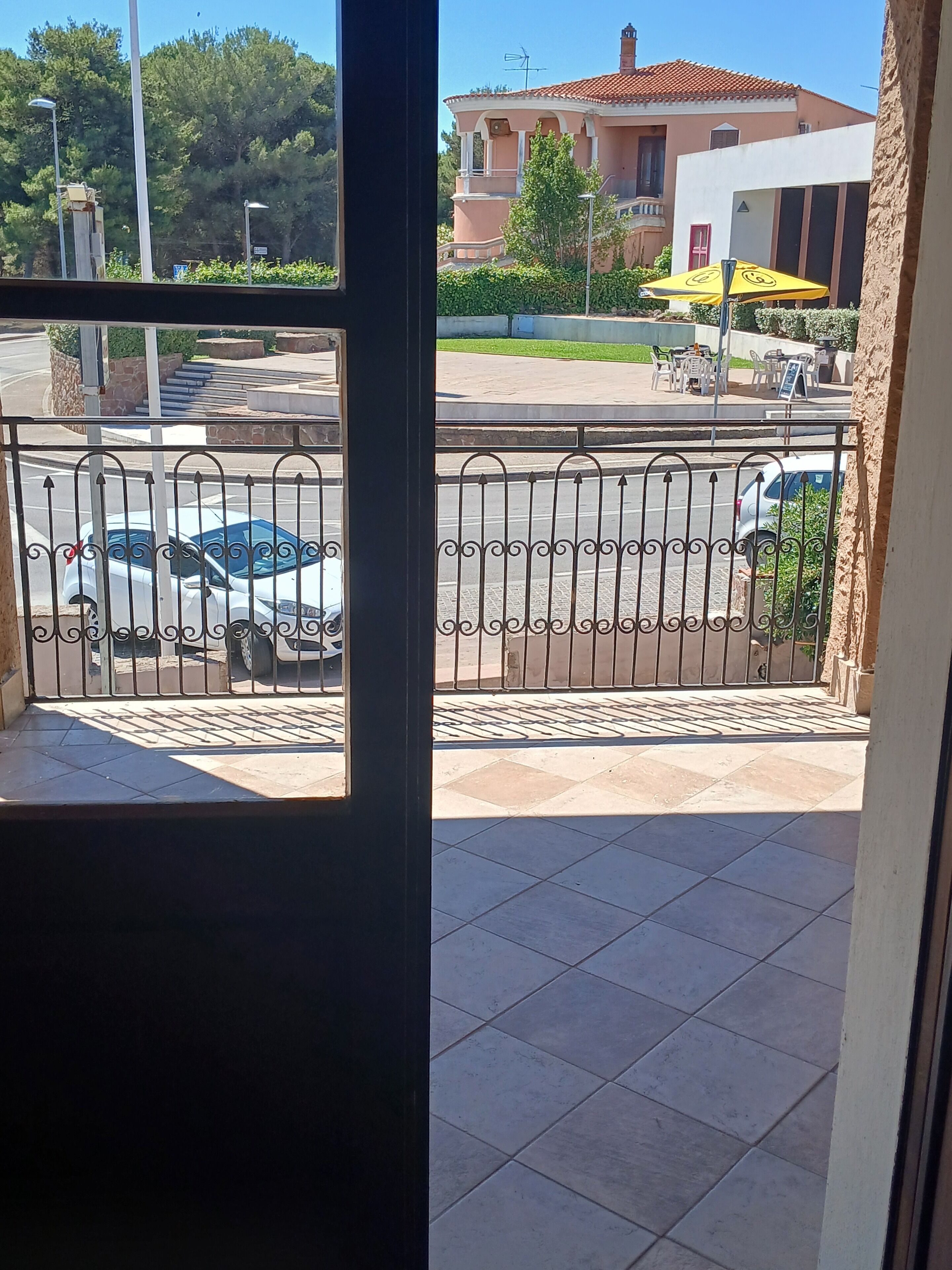 A balcony with a metal railing, a tiled floor, and a view of parked cars and a building with a red roof.
