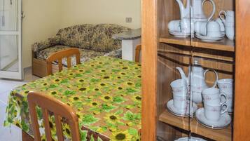 Un coin repas avec une table recouverte d’une nappe à motifs de tournesols, une chaise en bois, un canapé et un buffet contenant des services à thé en céramique blanche.