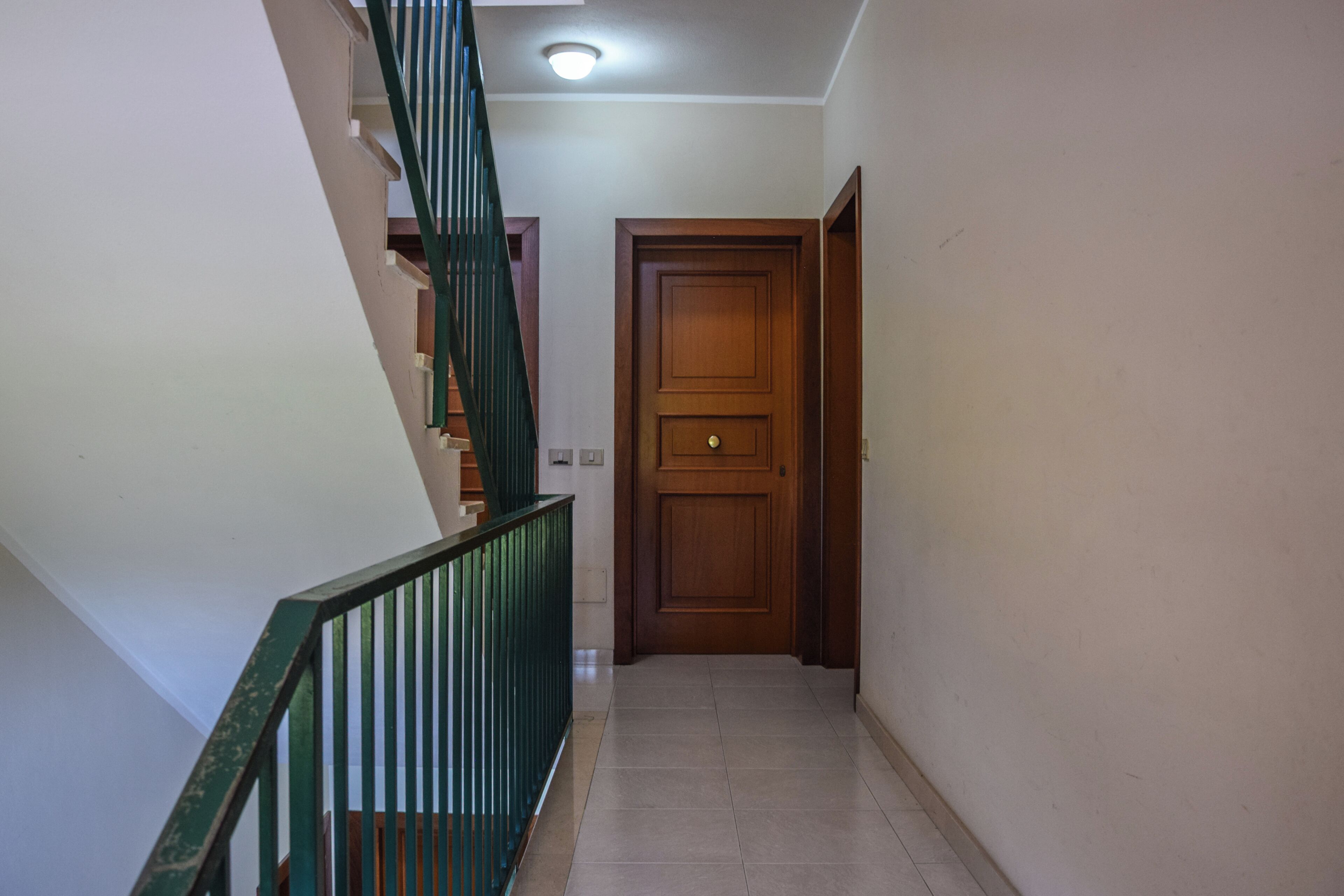 A hallway with a wooden door, a staircase with green railings, and tiled flooring.