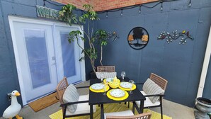 En patio med et bord dækket til fire personer, omgivet af fletstole, en potteplante og en dekorativ vægdekoration.