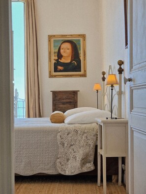 Une chambre à coucher avec un lit, une table de chevet avec des lampes, une commode en bois, un tableau encadré au mur et une fenêtre avec des rideaux.