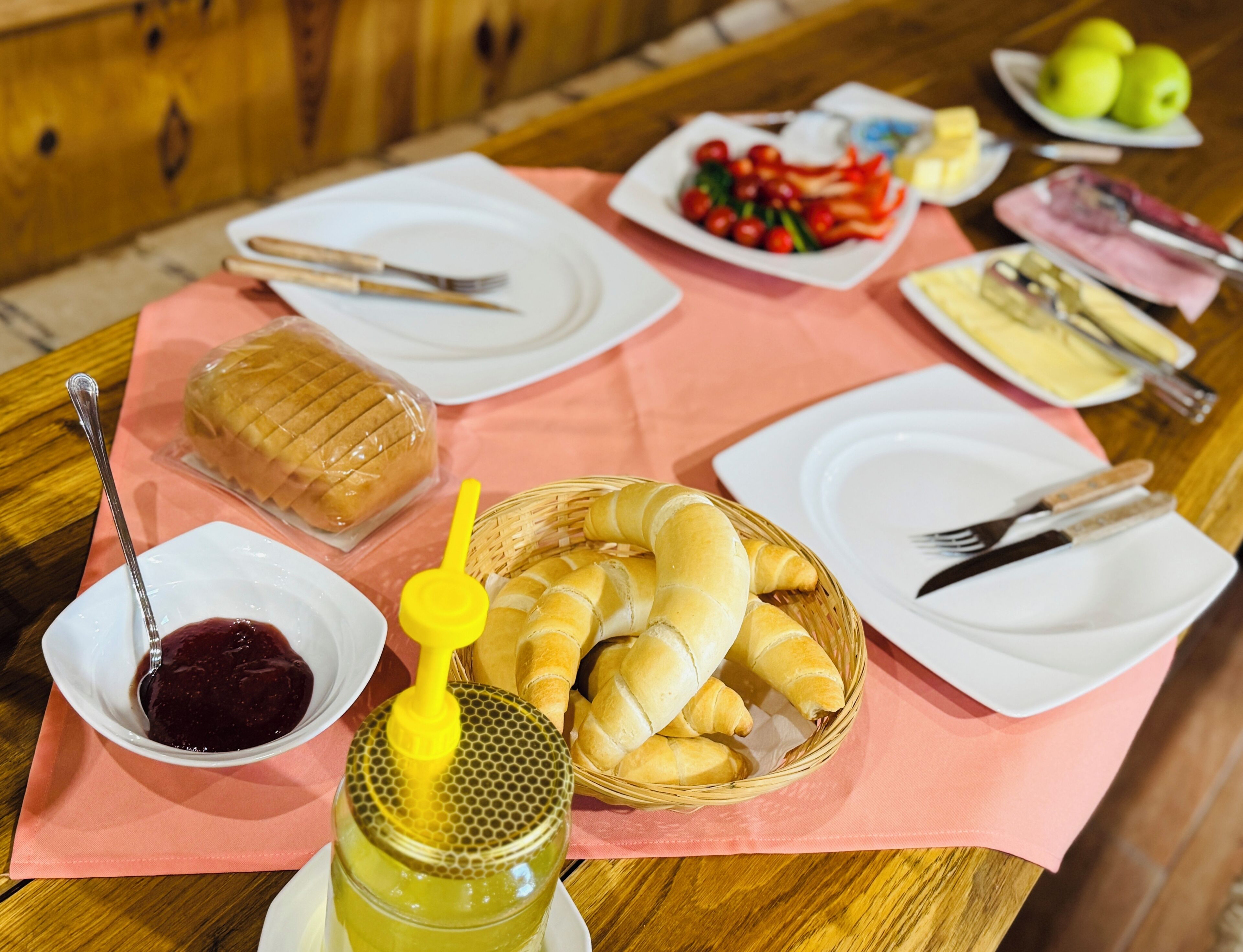 Een tafel met eten, waaronder brood, een mandje croissants, een pot honing en een schaal fruit.