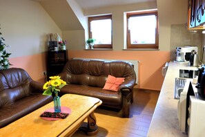 A living room with a brown leather sofa, a wooden coffee table, and a vase with flowers. There is a kitchen area with a coffee machine and a window with blinds.