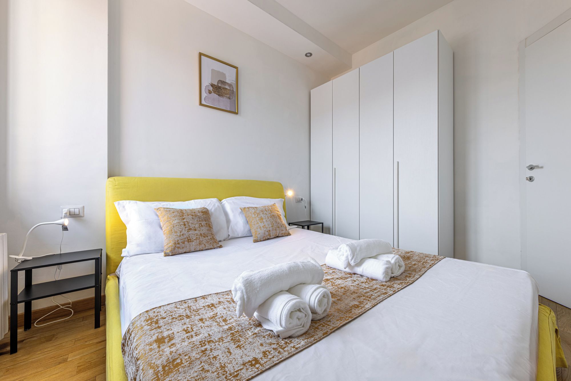 A hotel room with a bed, a bedside table, a wardrobe, and a wall-mounted picture.