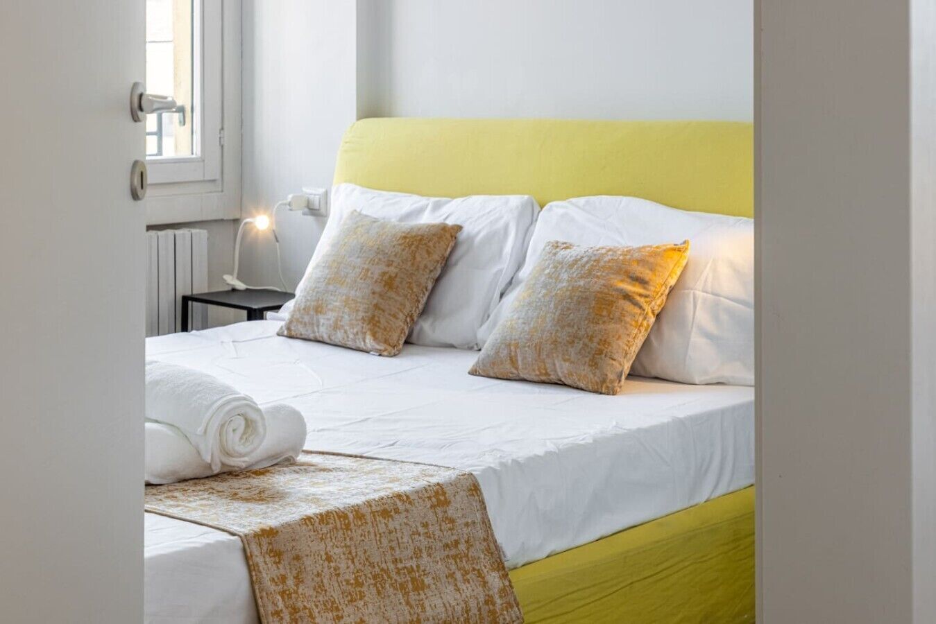 Un lit bien fait, avec du linge de lit blanc, deux oreillers à motifs et une tête de lit jaune.