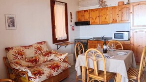 A compact living space with a sofa, dining table, chairs, kitchen area with appliances, and a window with curtains. - Casa Sunshine-Sea View (Costa Rei)