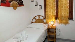 A single bed with white bedding, a bedside lamp, a wall-mounted fan, and a window with patterned curtains. - Casa Sunshine-Sea View (Costa Rei)