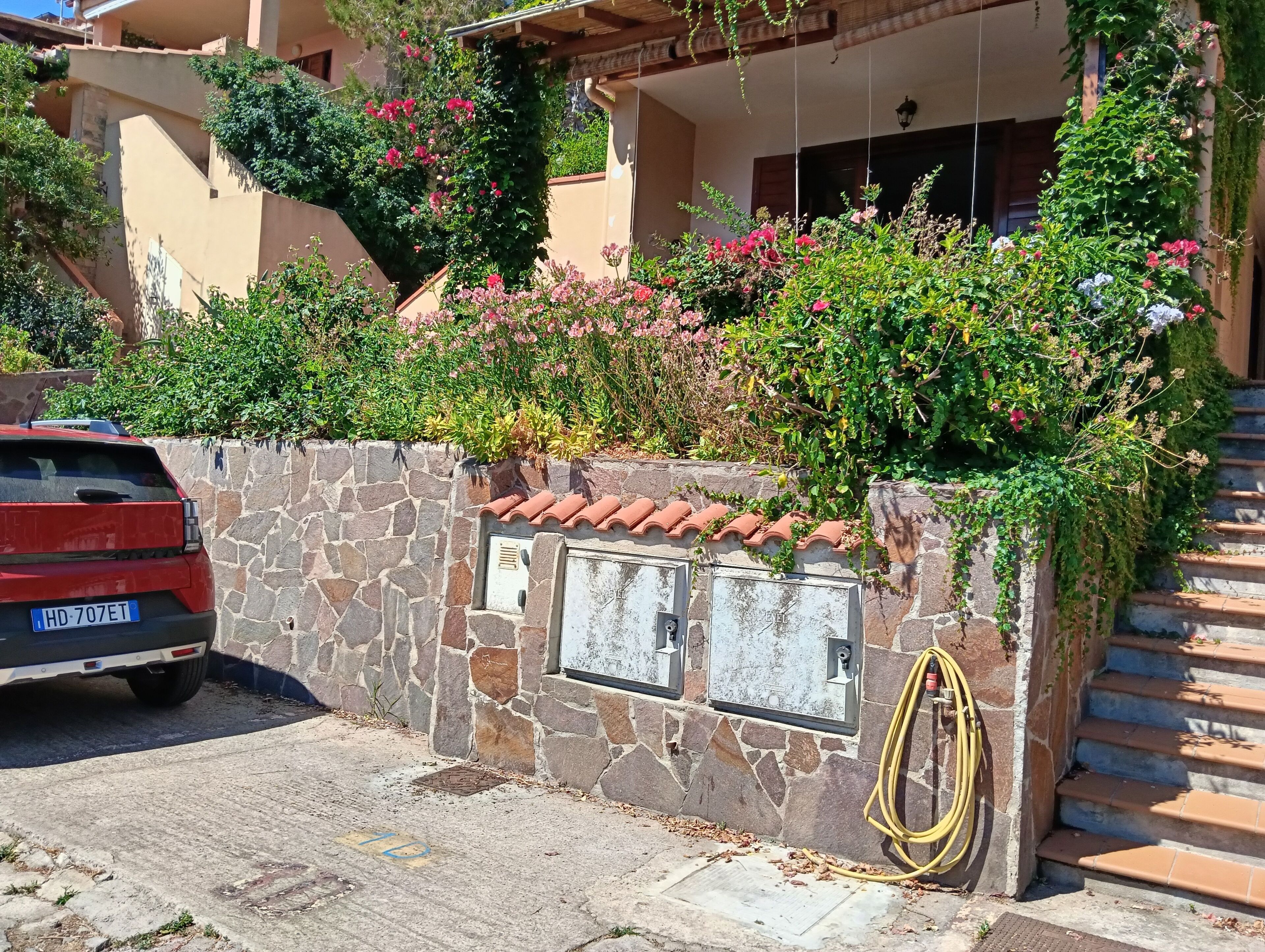 Un muro di pietra con un'auto parcheggiata davanti, un tubo da giardino e un edificio con tetto in tegole e piante rampicanti.