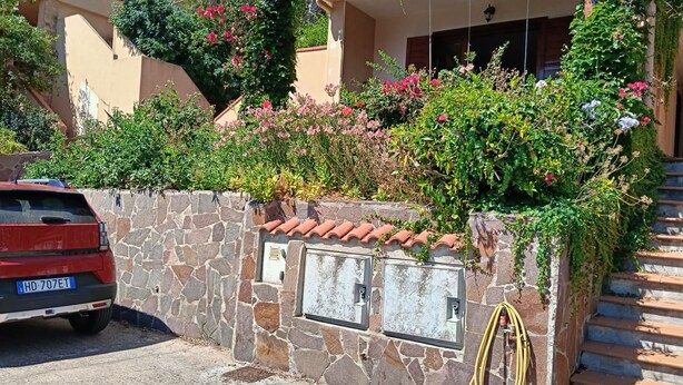 A stone wall with a car parked in front, a hose, and a building with a tiled roof and plants growing on it. - Casa Sunshine-Sea View (Costa Rei)