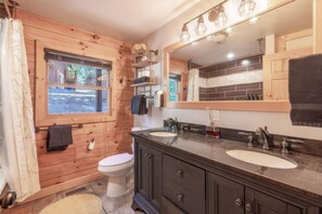 Een houten badkamer in een vakantiehuisje, met toilet, wastafelmeubel met twee wasbakken, een grote spiegel en een raam met uitzicht op de natuur.