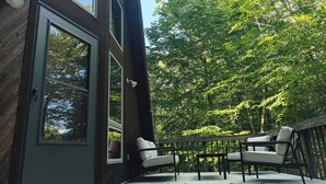 A modern wooden building with large windows, a deck with outdoor furniture, and a lush green tree backdrop.