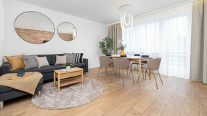 Una sala de estar moderna con sofá, mesa de centro, mesa de comedor, sillas y ventanas amplias con cortinas.
