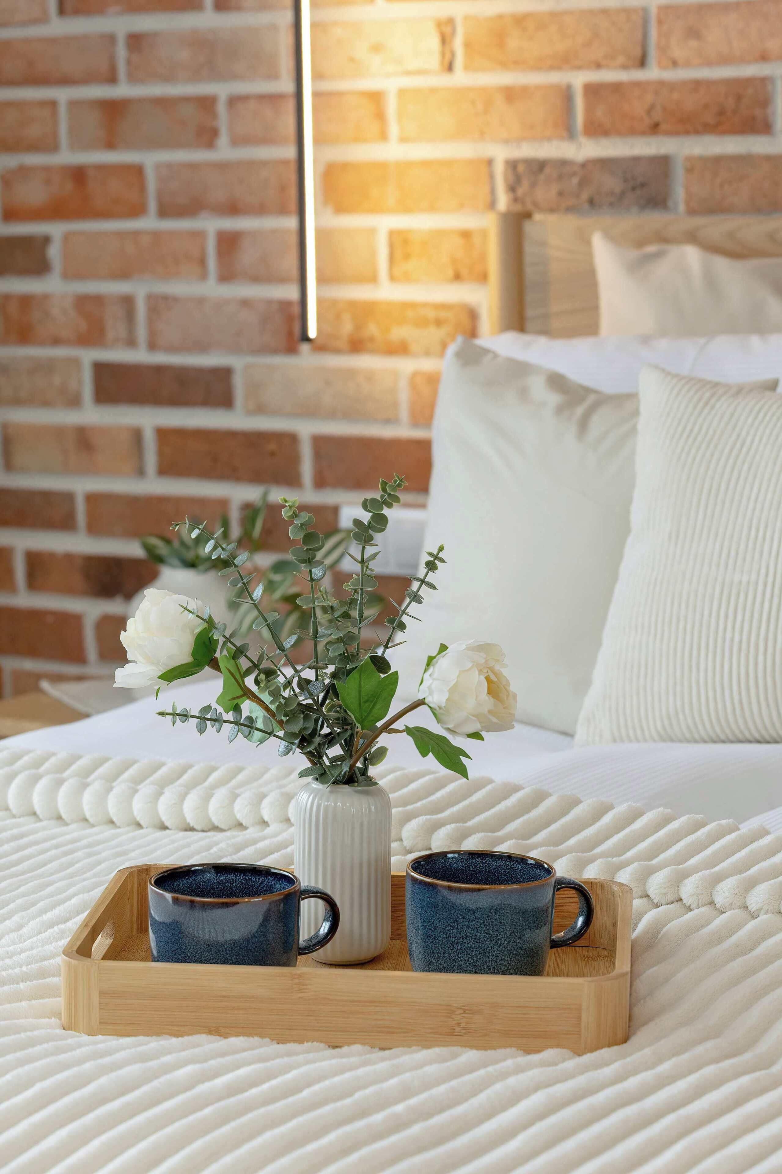 A bed with a white textured blanket, a wooden tray with two ceramic mugs, and a vase with white flowers on a brick wall background.