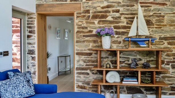 Um quarto com parede de pedra, um sofá azul, uma prateleira de madeira com objetos decorativos e uma maquete de veleiro.