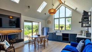 Uma sala de estar moderna com lareira, área de jantar, cozinha com armários azuis e janelas amplas com vista para o verde.