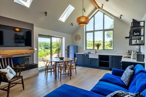 Ein modernes Wohnzimmer mit Kamin, Essbereich und Küche mit blauen Schränken, großes Fenster mit Blick auf Grünflächen.