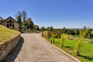 A gravel driveway leading to a wooden building with a sloped roof, surrounded by a grassy area and a wooden fence. - The Cart Shed (St Issey)